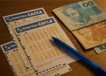 Bilhetes das Loterias da Caixa em cima de uma mesa. Há uma caneta em cima do bilhete e ao lado algumas notas