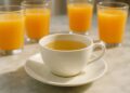 Uma xícara de chá branco, ao fundo 4 copos de suco de laranja