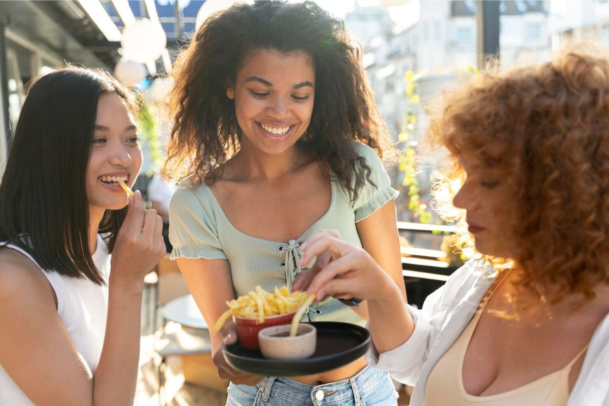 Um trio de amigas comendo e vendo Como economizar com alimentação fora de casa