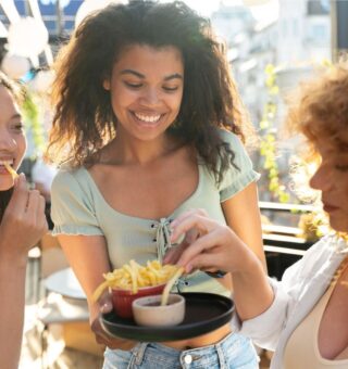 Um trio de amigas comendo e vendo Como economizar com alimentação fora de casa